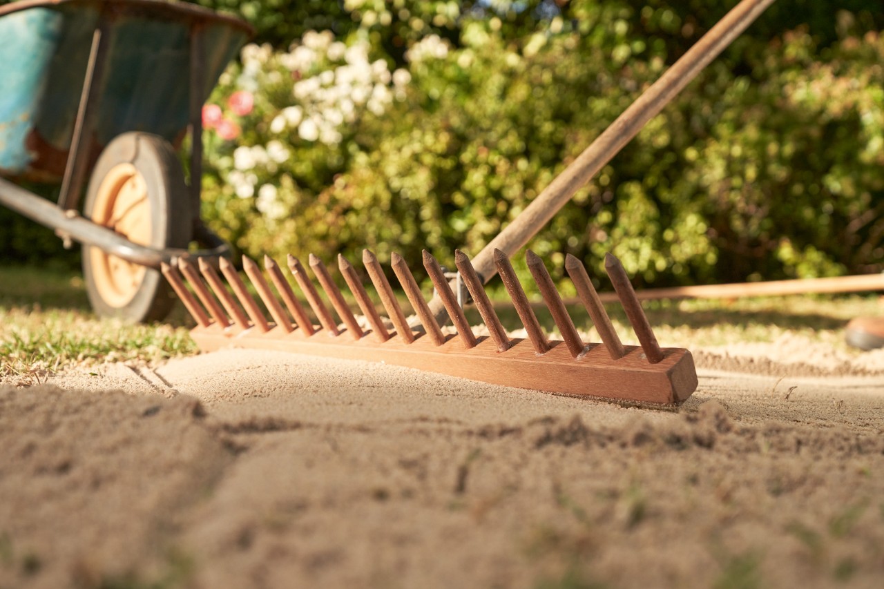 Met de achterkant van een hark wordt zand op een vlakke ondergrond geëgaliseerd, op de achtergrond kruiwagen en heg
