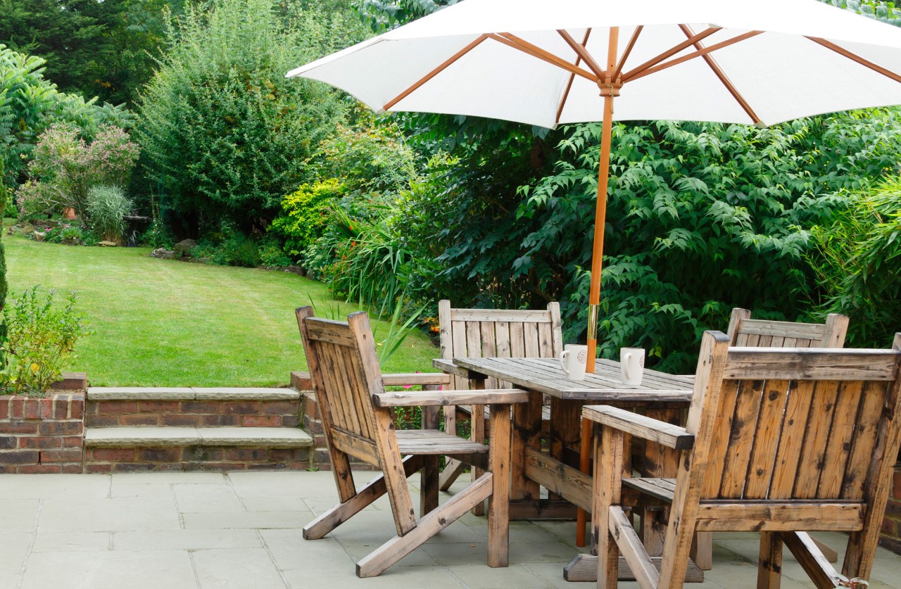 Verlaagd terras in een kleine tuin met tuinmeubels op de voorgrond, gazon met struiken en kleine bomen erachter.