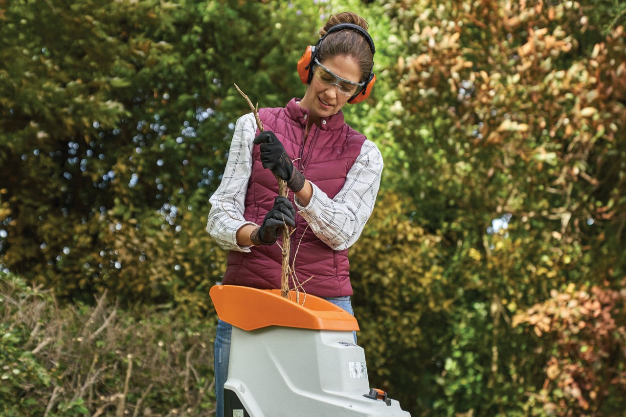 Een vrouw draagt handschoenen, gehoorbescherming en een bril terwijl ze takken in de hakselaar stopt