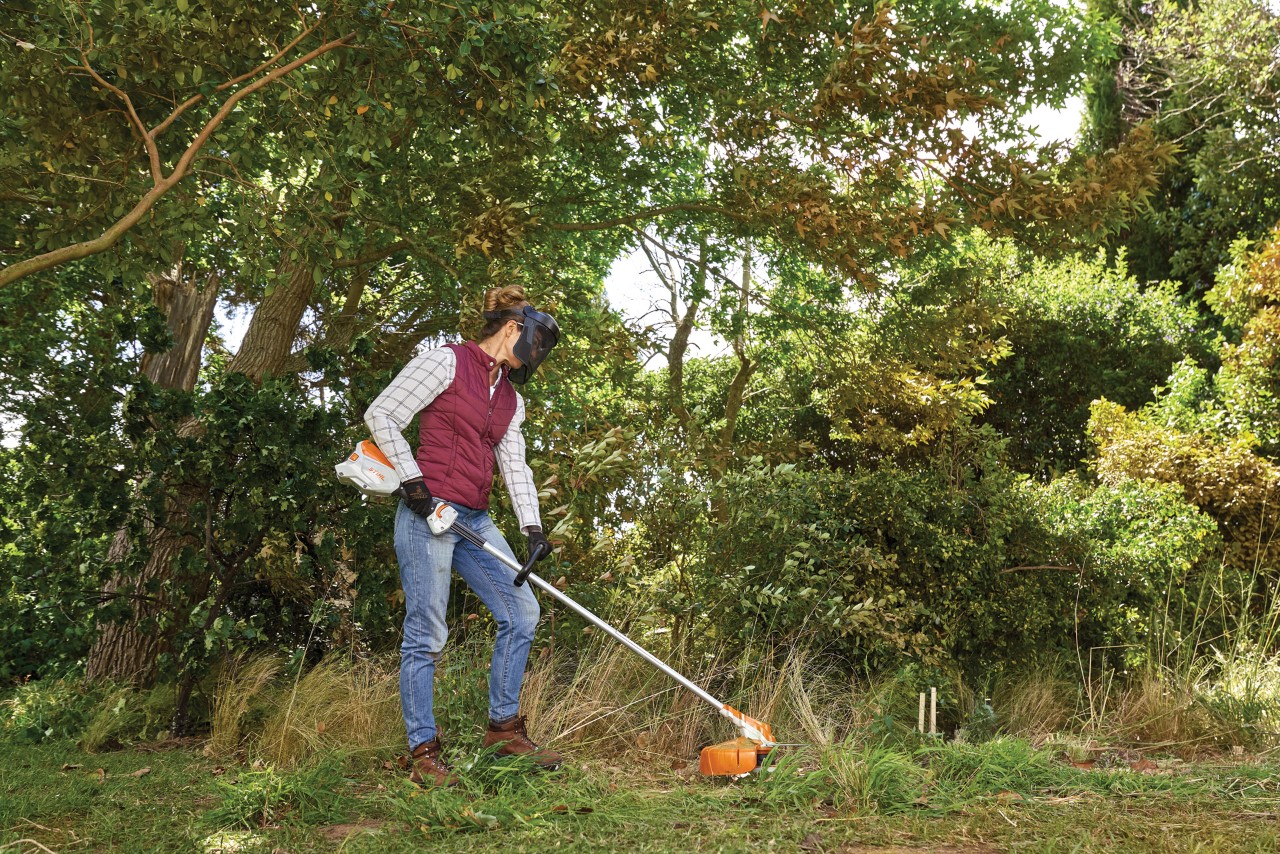 Een vrouw met bosmaaier voor overwoekerd gras met bomen en struiken op de achtergrond