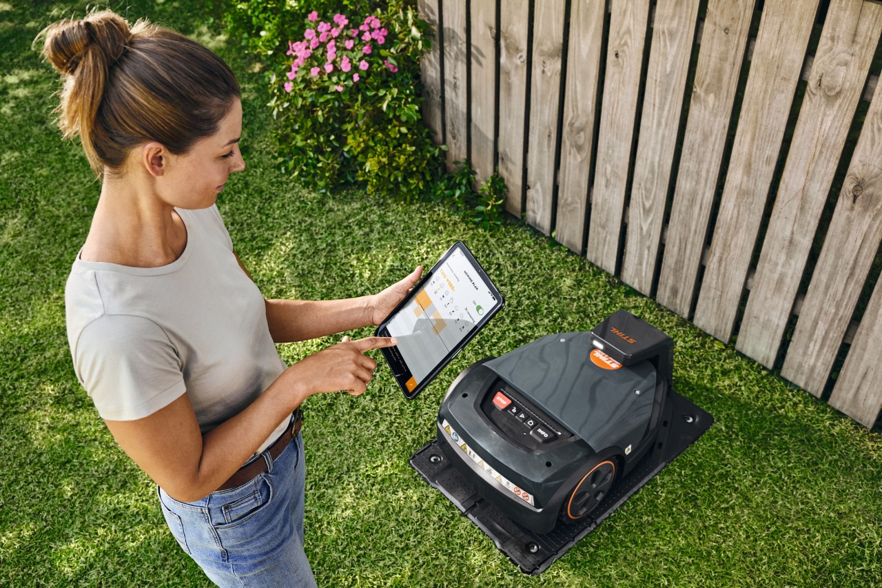 Een vrouw staat voor een STIHL iMOW® robotmaaier op het gazon en houdt een tablet met de geopende iMOW® app in haar hand 